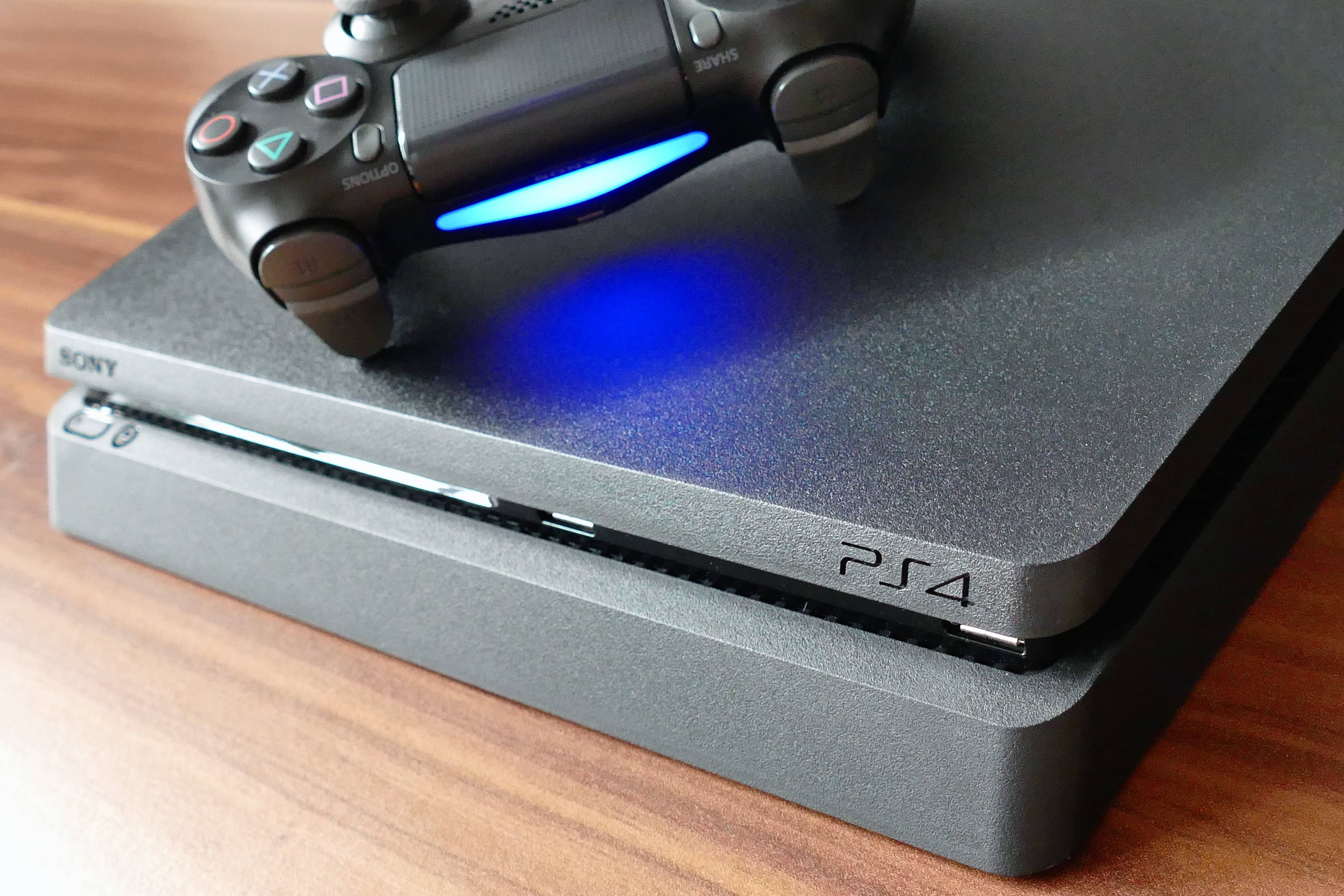 Best Ps4 桌布 壁紙 — 黑色PlayStation 4主機的特寫，上面有一個發出藍光的控制器，放在木質表面上。