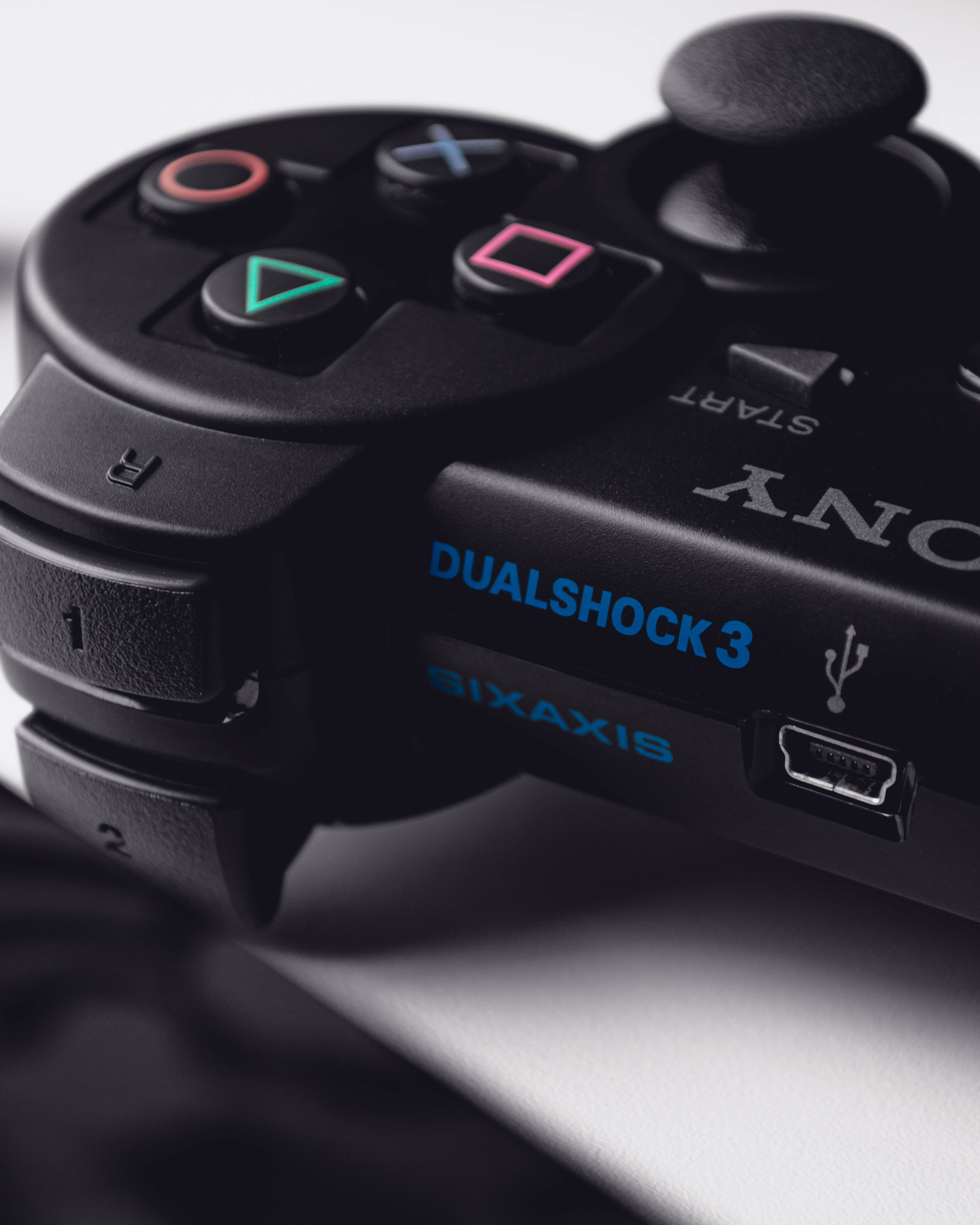 PS3 桌布 壁紙 — 黑色PlayStation DualShock 3手把的特寫，帶有發光的彩色按鈕和藍色文字，置於柔軟的白色表面上。