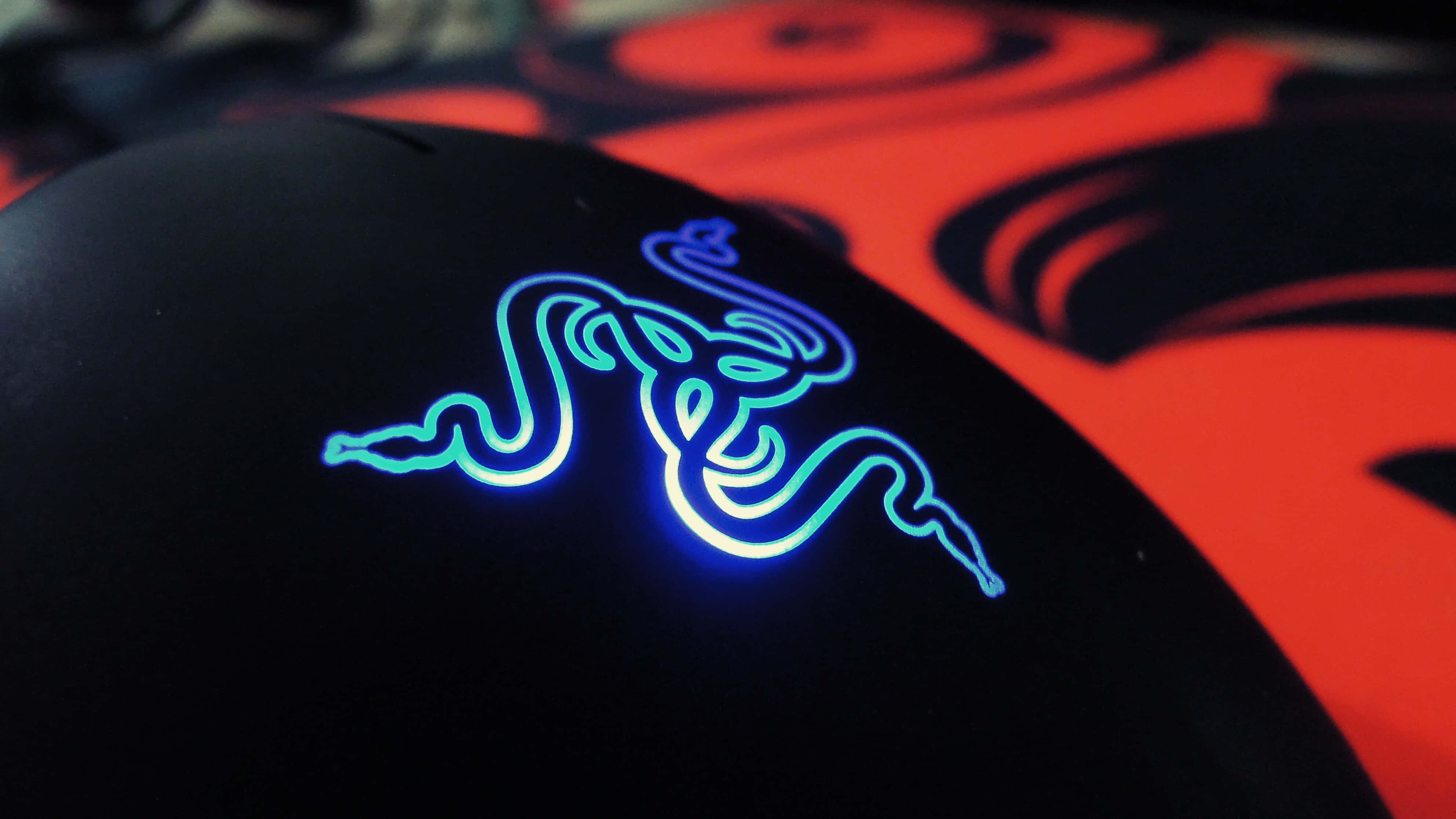 Gaming Mice 桌布 壁紙 — 黑色Razer滑鼠特寫，帶有發光藍色標誌，背景為紅色抽象圖案。