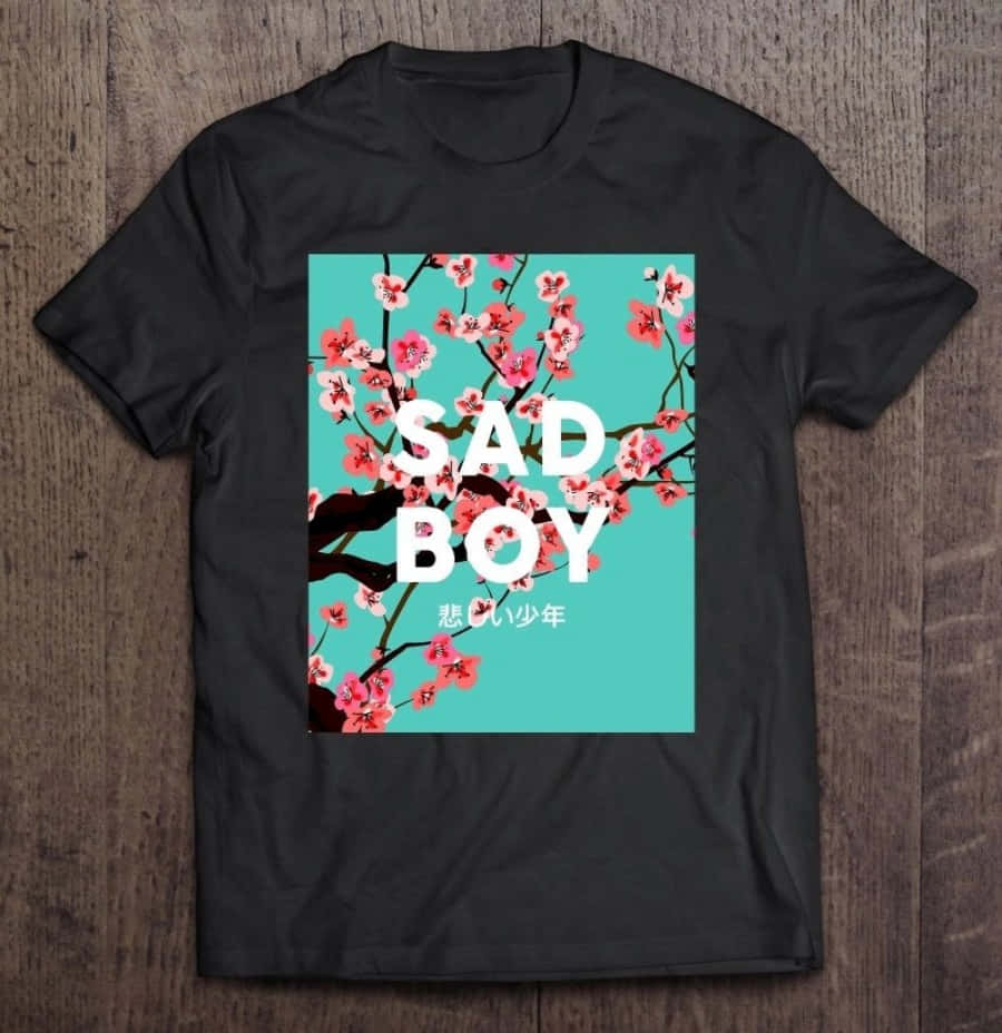 T Shirt 桌布 壁紙 — 黑色T恤，白色粗體「SAD BOY」文字和粉紅色櫻花，青色背景，放在木質表面上。