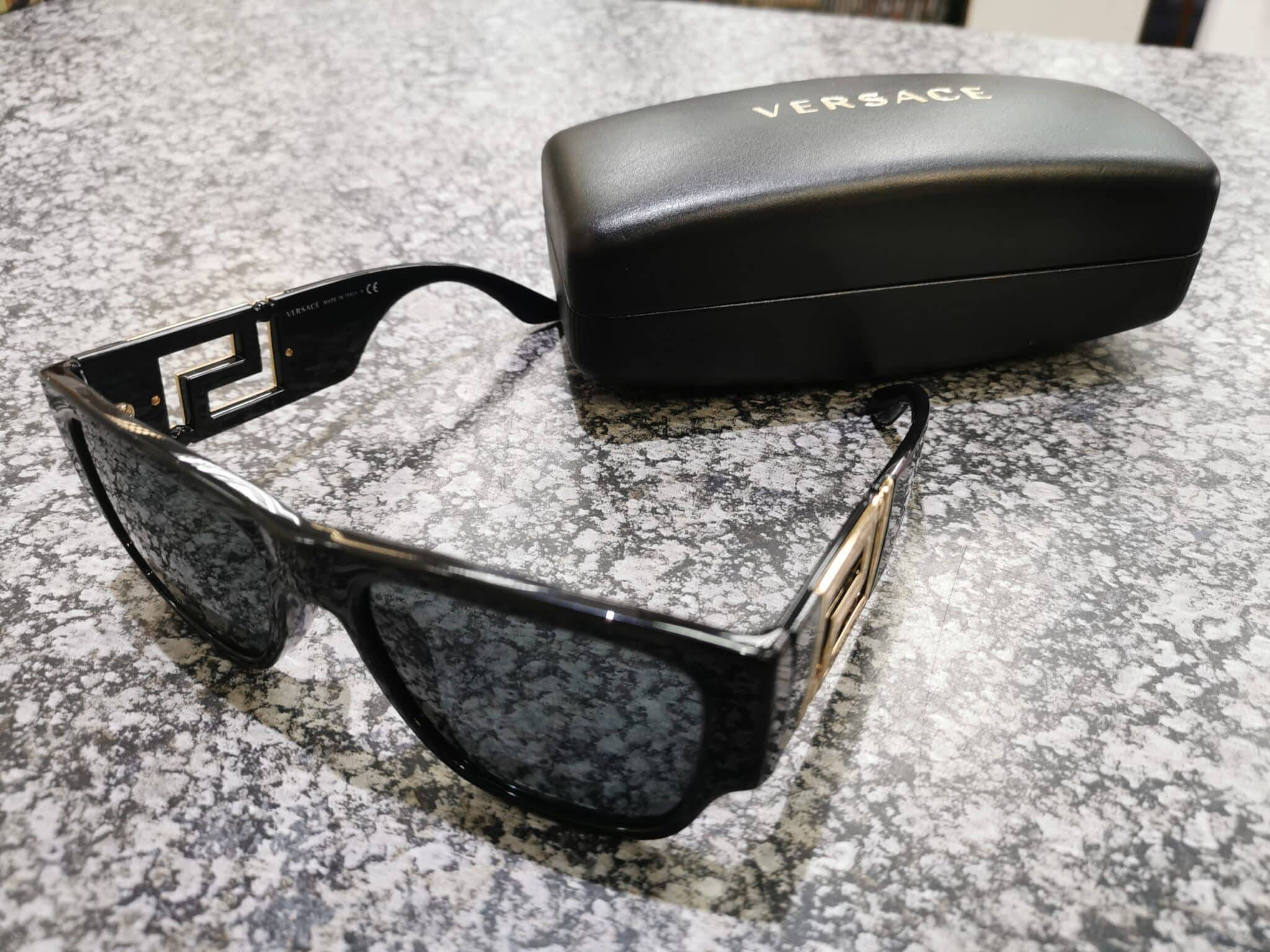 Sunglasses 桌布 壁紙 — 黑色Versace太陽眼鏡帶金色標誌細節，旁邊是其品牌眼鏡盒，放在斑點灰色檯面上。