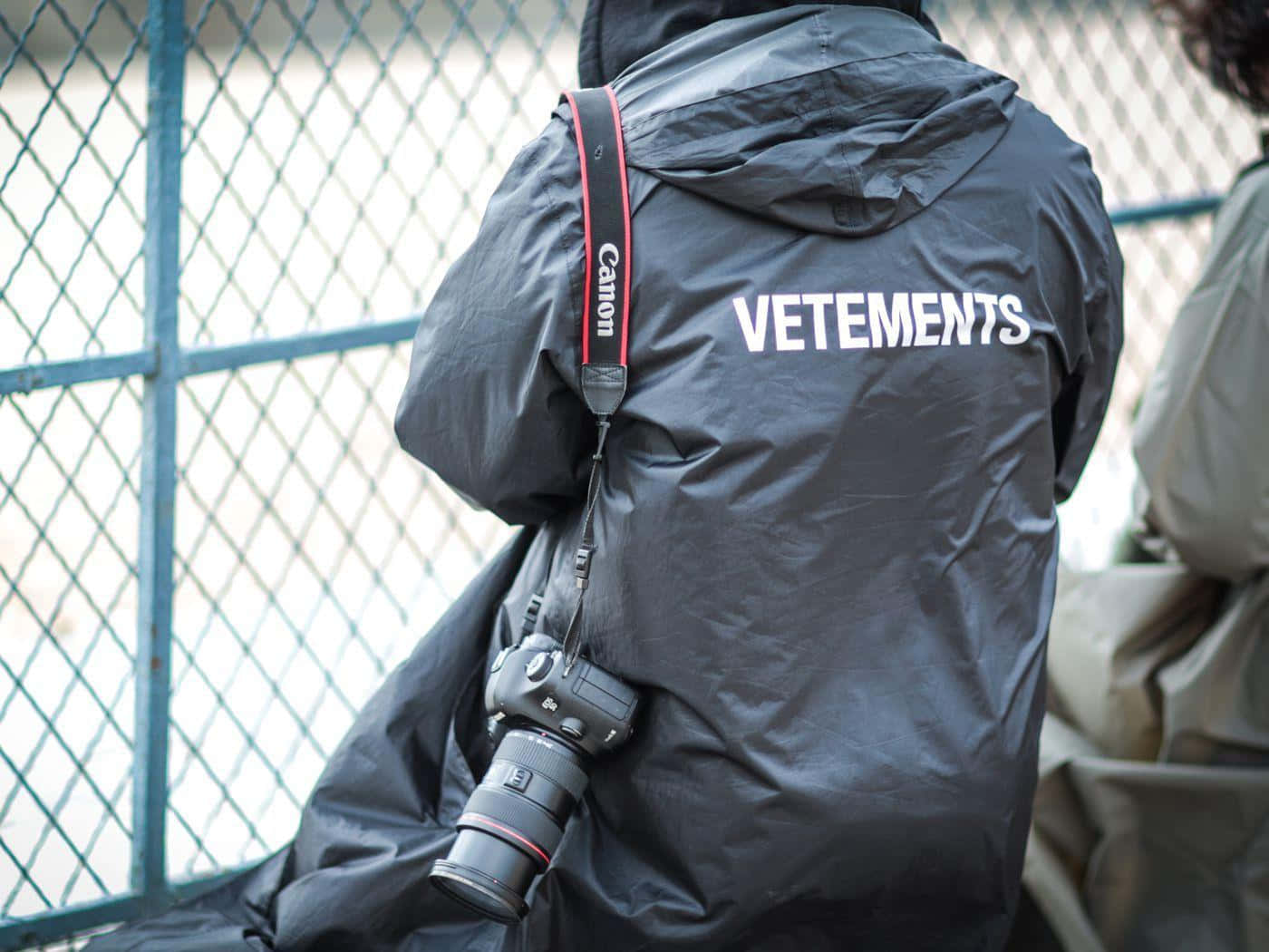 vetements 桌布 壁紙 — 黑色Vetements外套人士背面，肩上掛著Canon相機，靠近鐵絲網圍欄。
