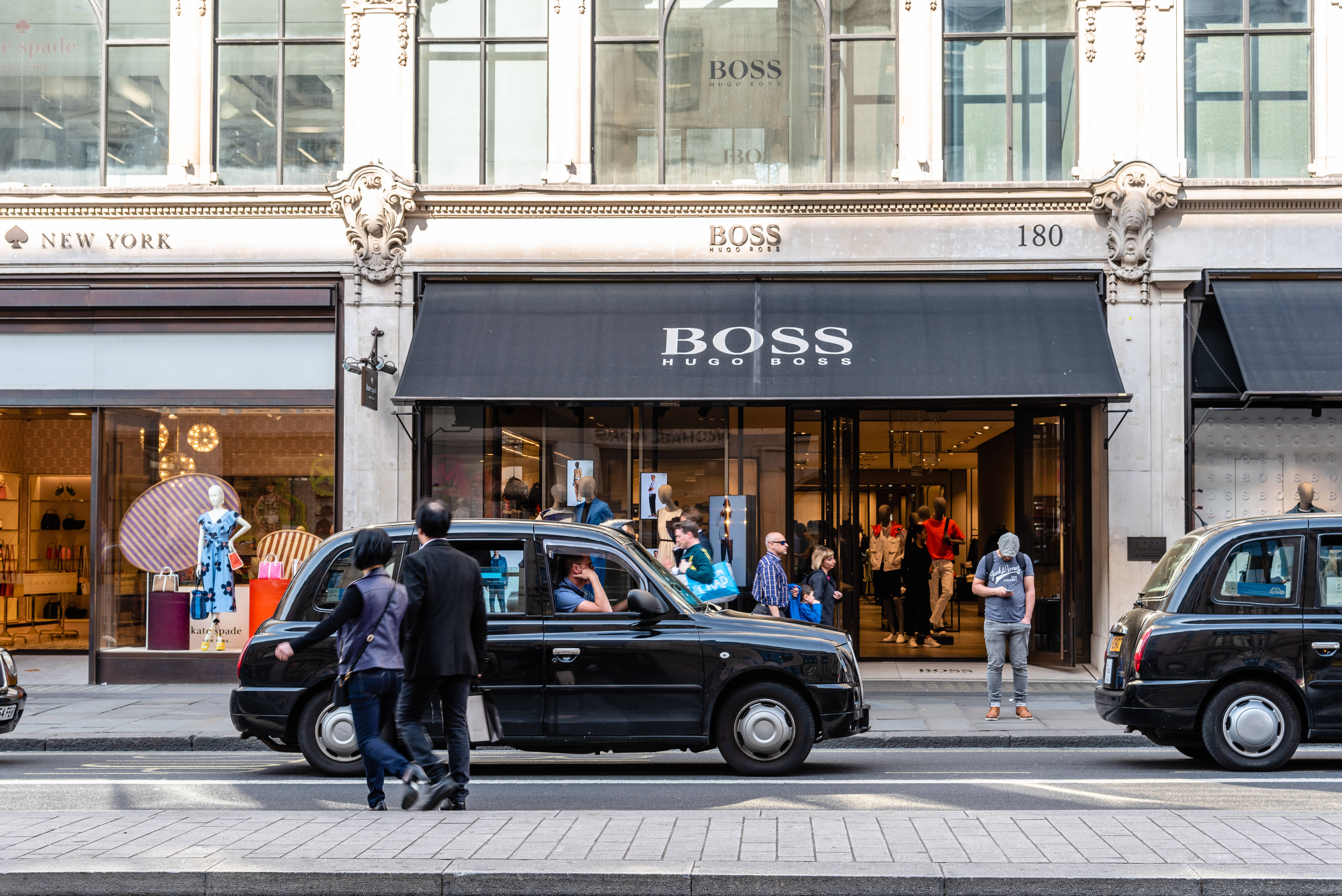 Building 桌布 壁紙 — 黑色倫敦計程車駛過Hugo Boss商店，行人在明亮日光下在人行道上行走。