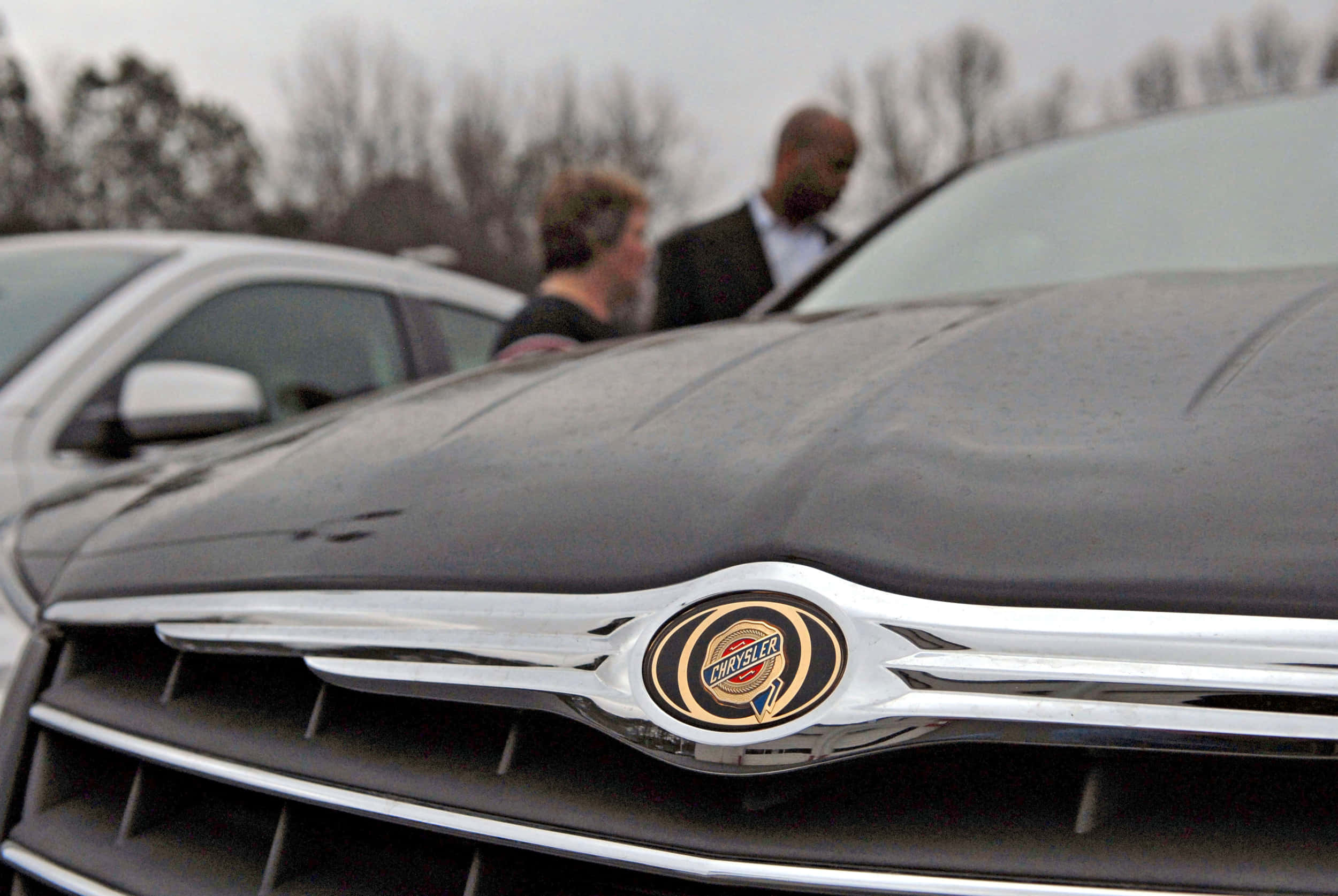 Chrysler 桌布 壁紙 — 黑色克萊斯勒格柵近距離特寫，帶有鍍鉻徽章，前景是模糊的人群和汽車，在陰天的戶外環境中。