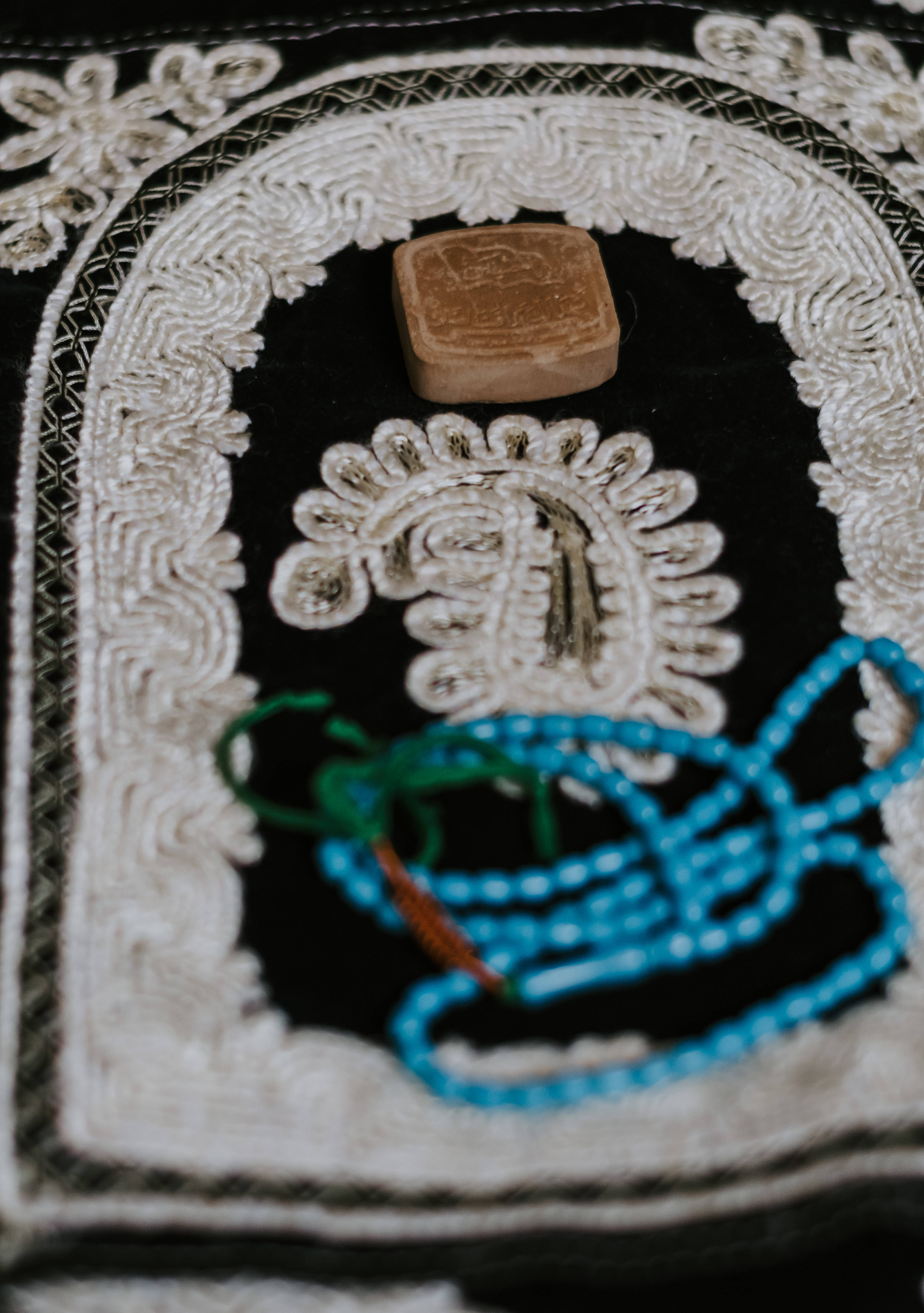 真主 桌布 壁紙 — 黑色刺繡布料近距離特寫，帶有白色花卉圖案，手持一個棕色方形物體和藍綠色念珠。