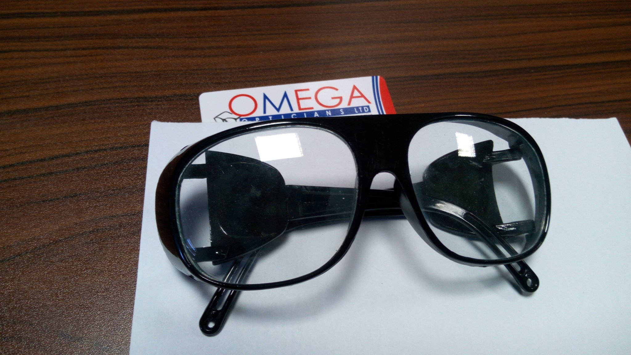 Sunglasses 桌布 壁紙 — 黑色厚框安全眼鏡放在木桌上的白紙上，旁邊有一張Omega Opticians卡片。