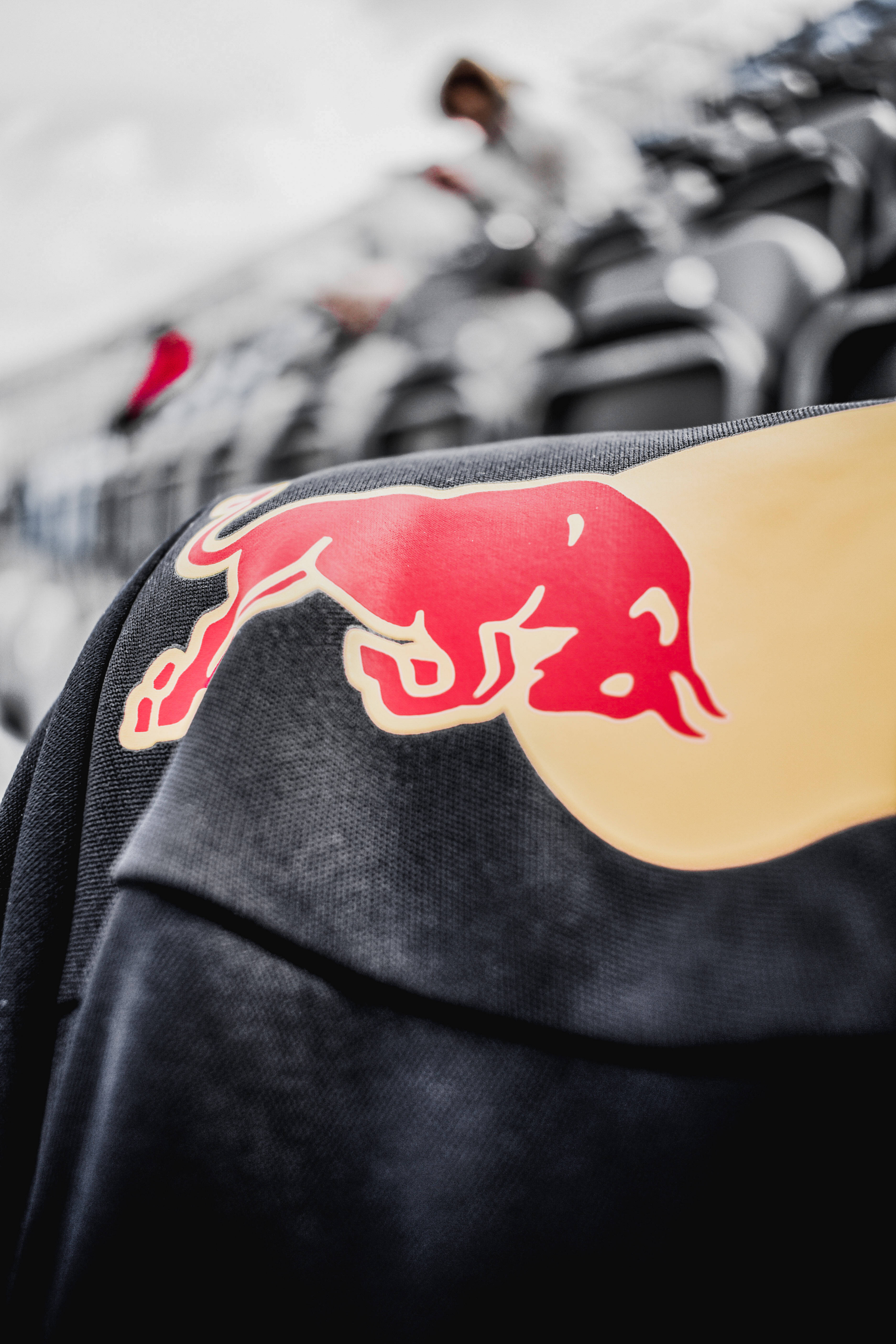 Red Bull 桌布 壁紙 — 黑色外套上紅色Red Bull標誌的特寫，背景為模糊的體育場座位。