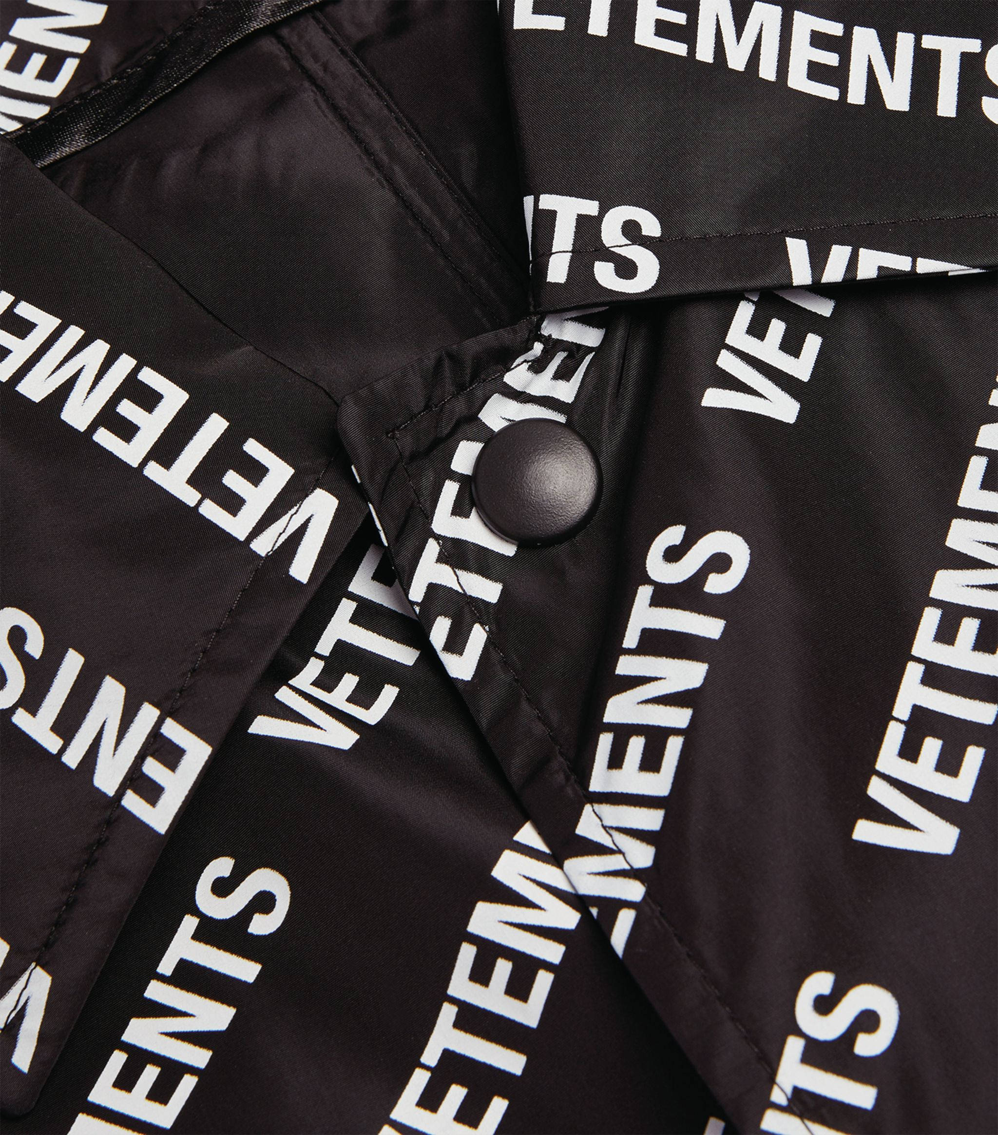 Vetements 桌布 壁紙 — 黑色外套的特寫，布料上對角重複印有粗體白色「VETEMENTS」文字，並有一顆黑色鈕扣。