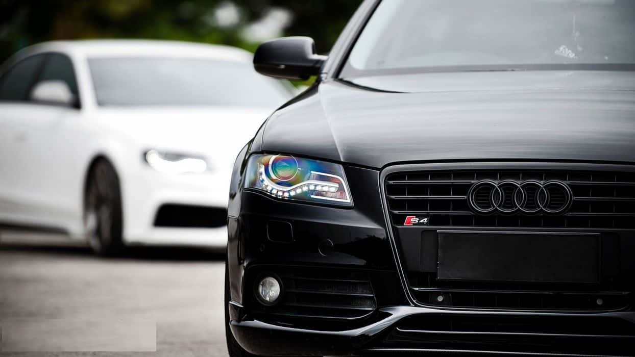Audi S4 桌布 壁紙 — 黑色奧迪S4的緊湊特寫，帶有發光的彩虹頭燈，背景中有一輛模糊的白色汽車。