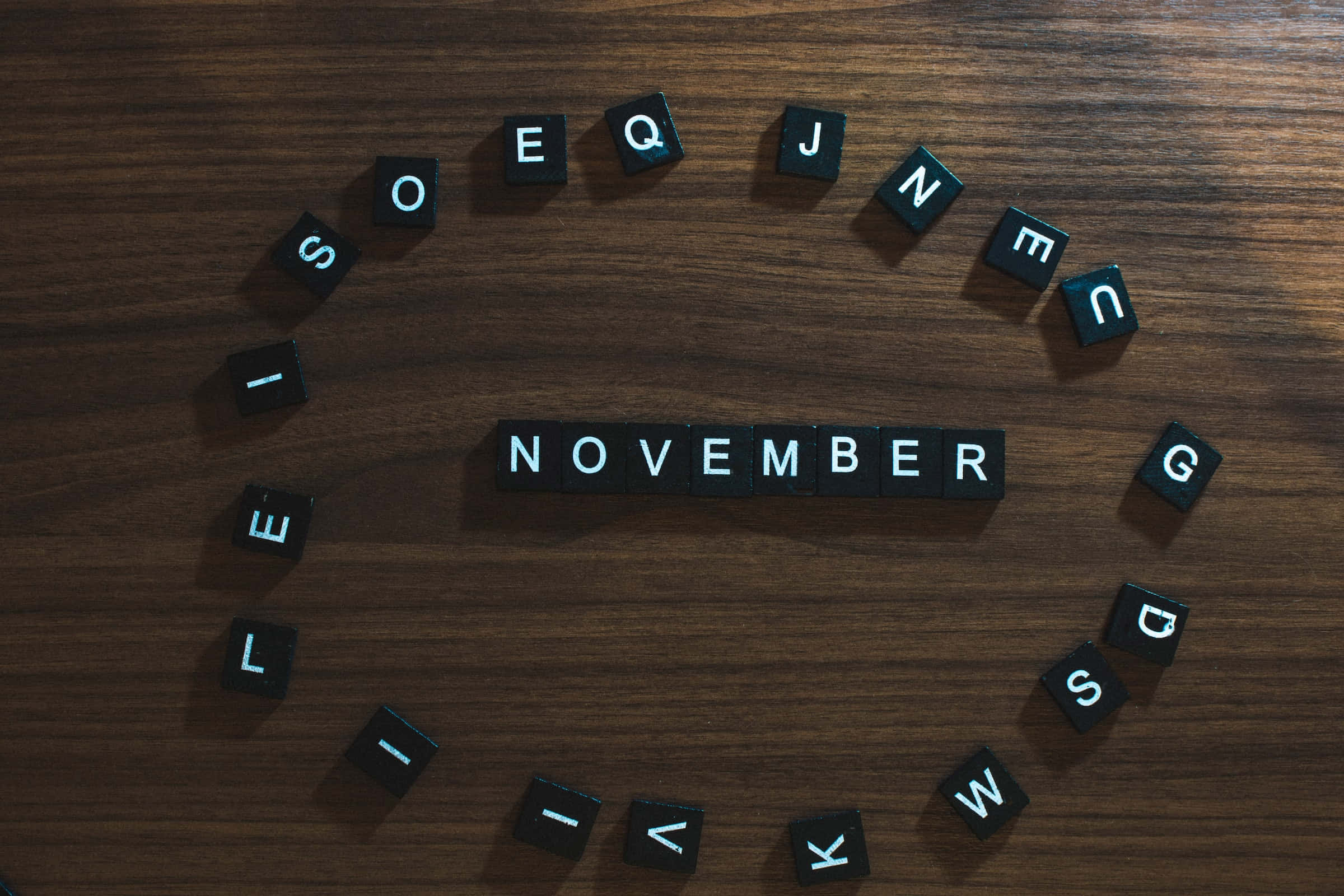 Cute November 桌布 壁紙 — 黑色字母瓷磚拼出「NOVEMBER」，居中放在深色木質表面上，周圍散落著字母瓷磚。