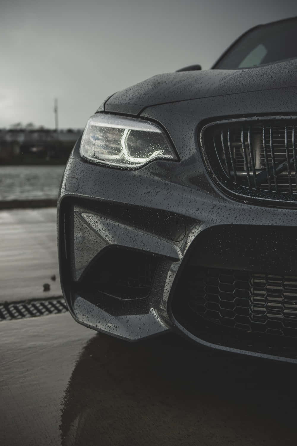Bmw Android 桌布 壁紙 — 黑色寶馬M2跑車前格柵和頭燈的特寫，在濕漉漉的路面上閃爍著雨滴。