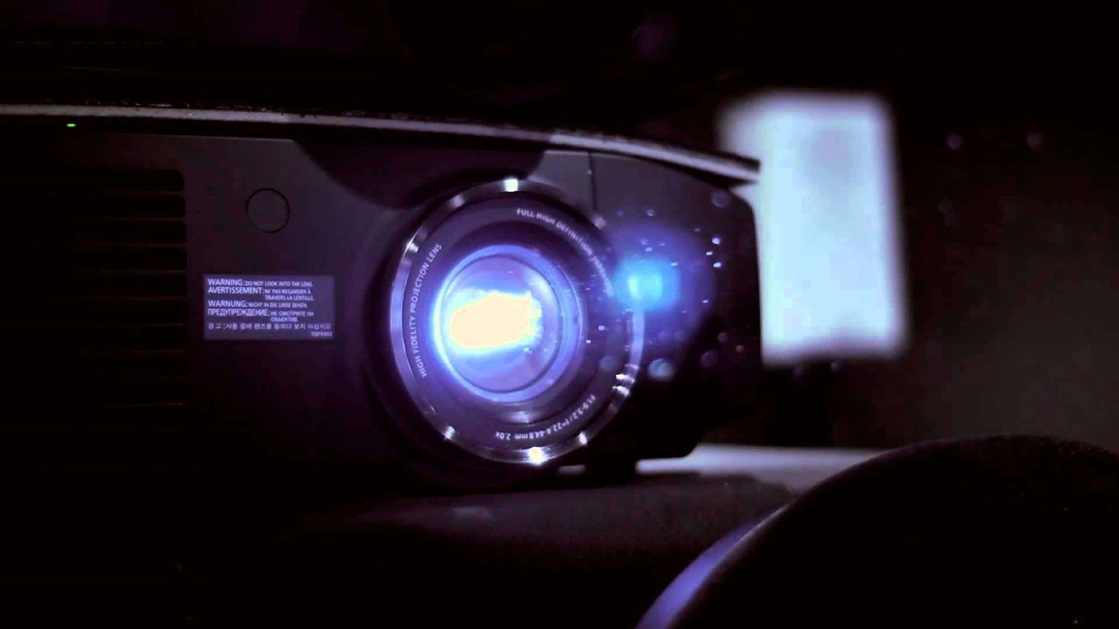 4K Projector 桌布 壁紙 — 黑色投影機在黑暗房間中發出藍光，在螢幕上投射出微弱圖像的特寫。
