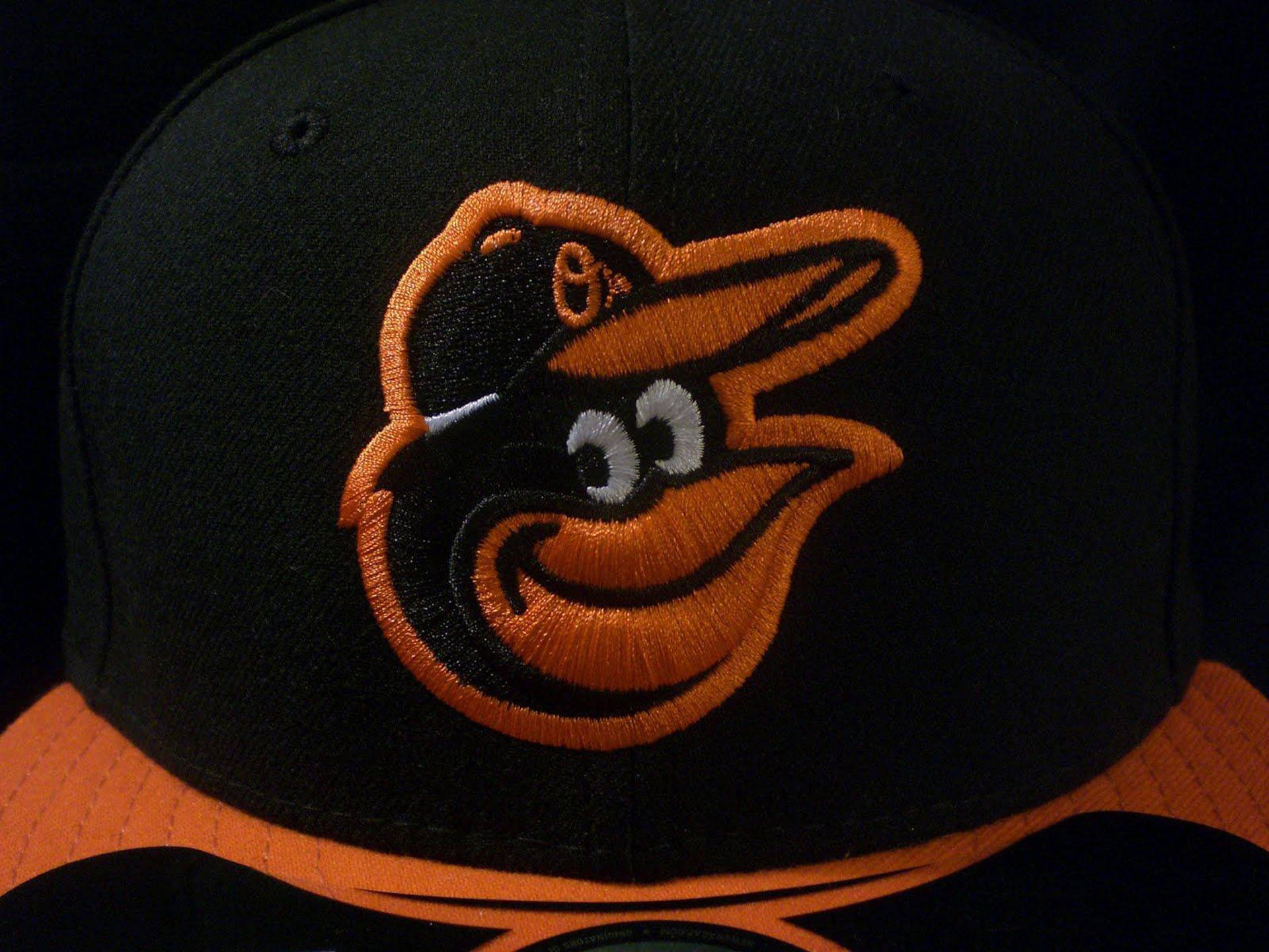 Orioles 桌布 壁紙 — 黑色棒球帽的特寫，帽上有一個橙色和黑色的刺繡黃鸝標誌，背景為深色。