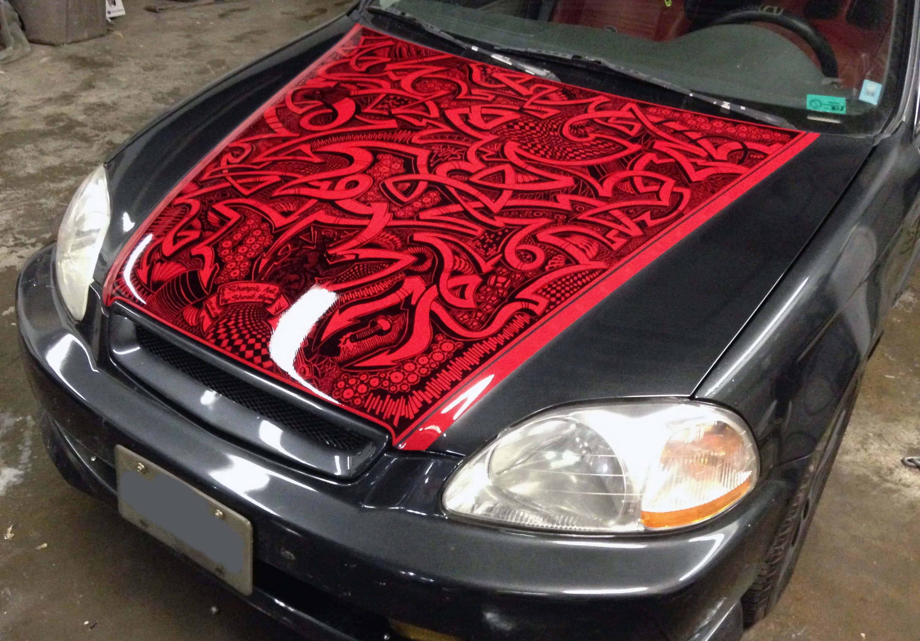 Car Hood 桌布 壁紙 — 黑色汽車的引擎蓋上有一個大膽的紅色部落風格噴繪設計，帶有複雜的漩渦圖案。