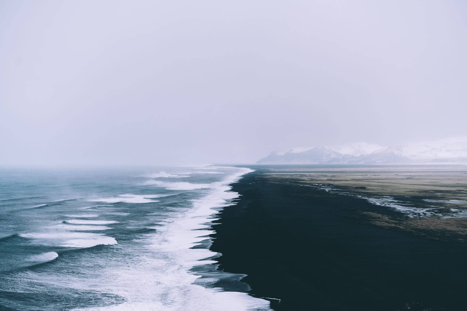 Sea 桌布 壁紙 — 黑色沙灘的清晰鳥瞰圖，海浪拍打著霧氣瀰漫的地平線，遠處有積雪的山峰。