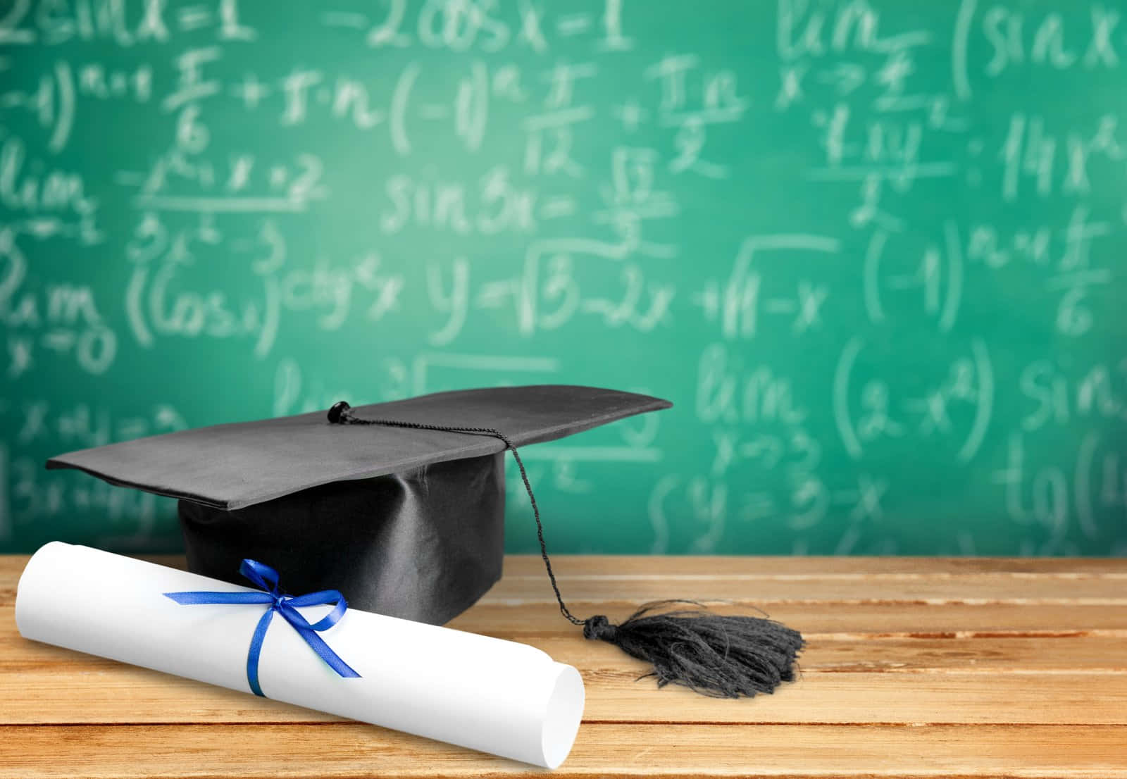 Graduation Ceremony 桌布 壁紙 — 黑色畢業帽和帶有藍色絲帶的畢業證書放在木桌上，前面是充滿數學方程式的綠色黑板。