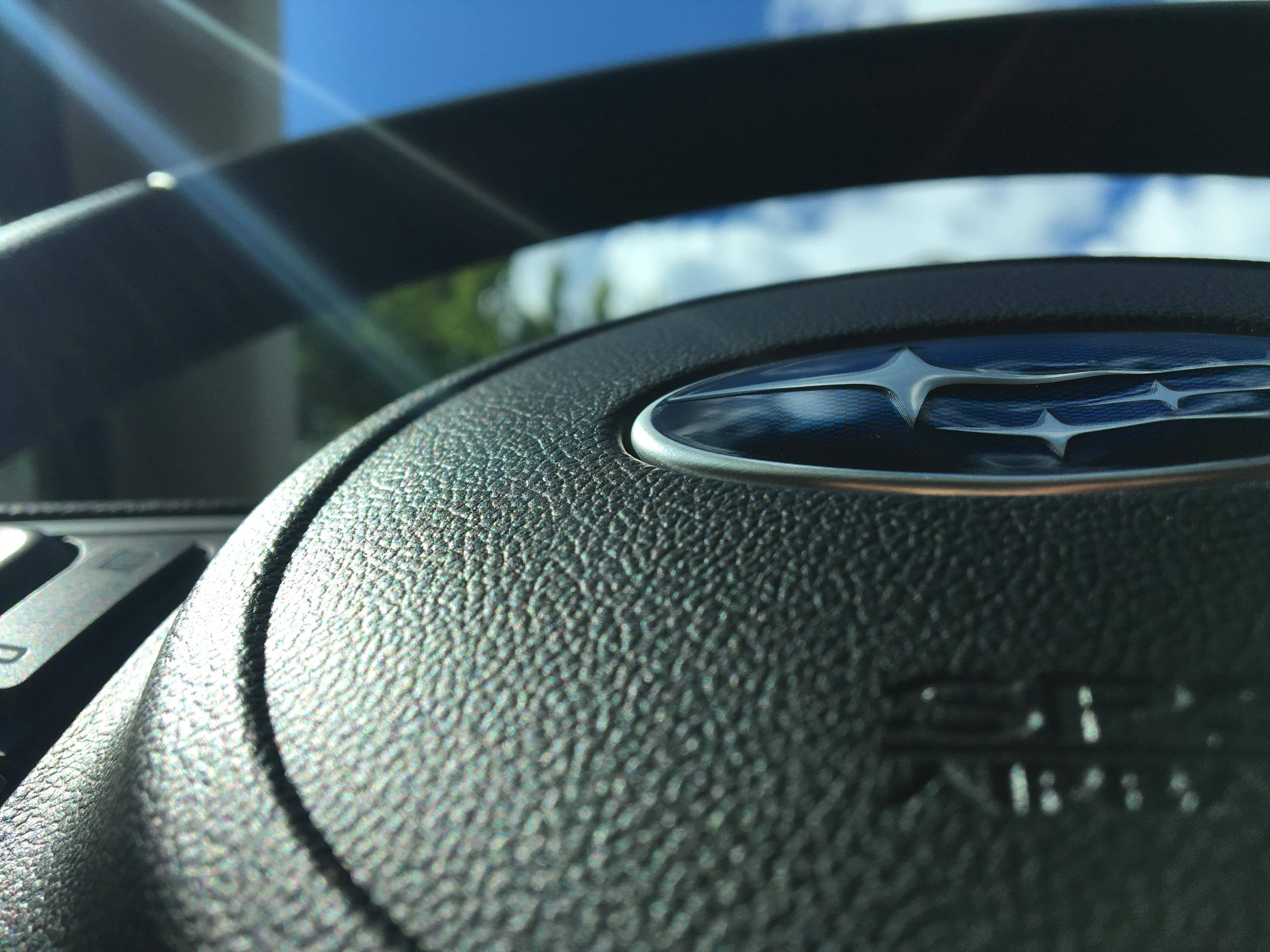 Subaru Logo 桌布 壁紙 — 黑色皮革 Subaru 方向盤連標誌在明亮陽光和藍天反射下的特寫。