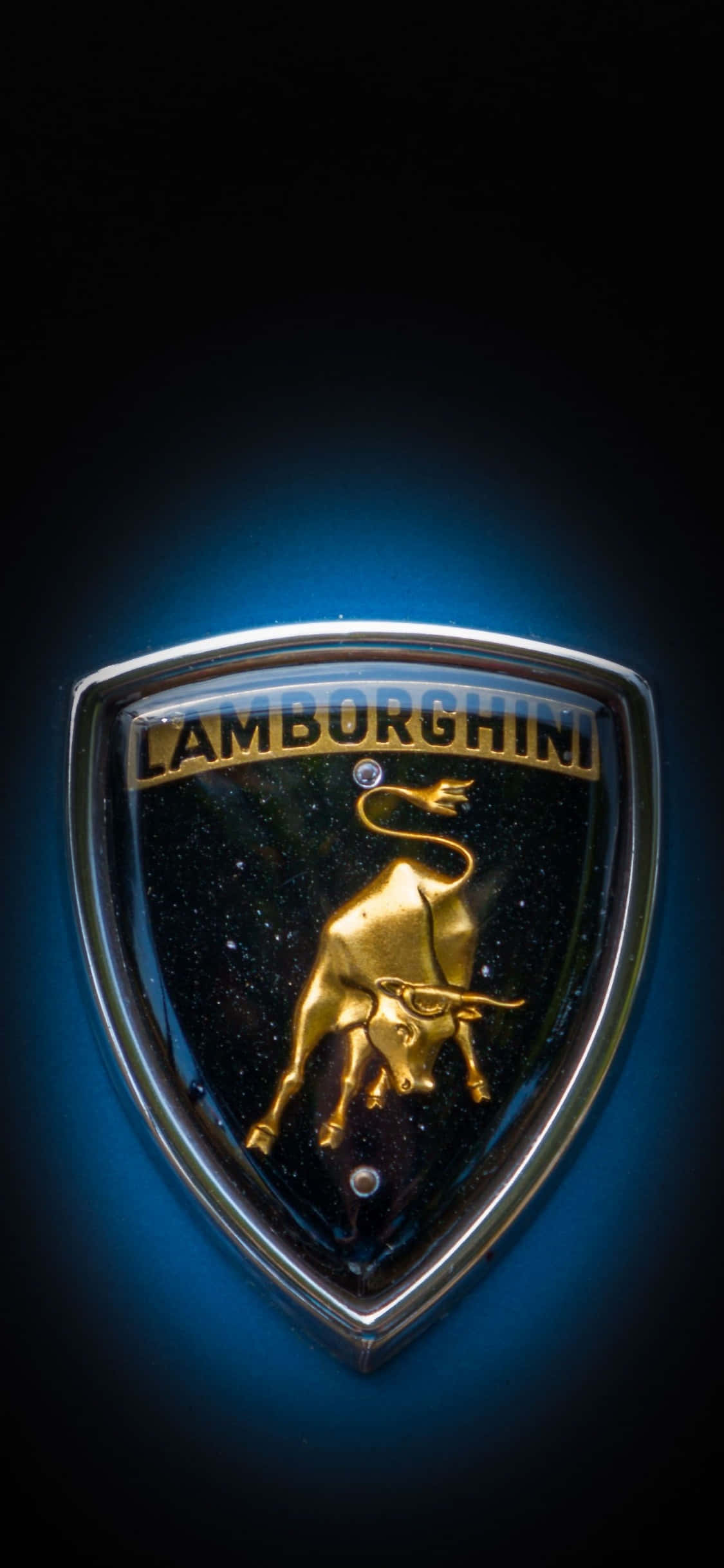 iPhone X Lamborghini 桌布 壁紙 — 黑色盾牌上閃閃發光的金色公牛標誌，帶有「LAMBORGHINI」文字，在深色背景下被藍光照亮。