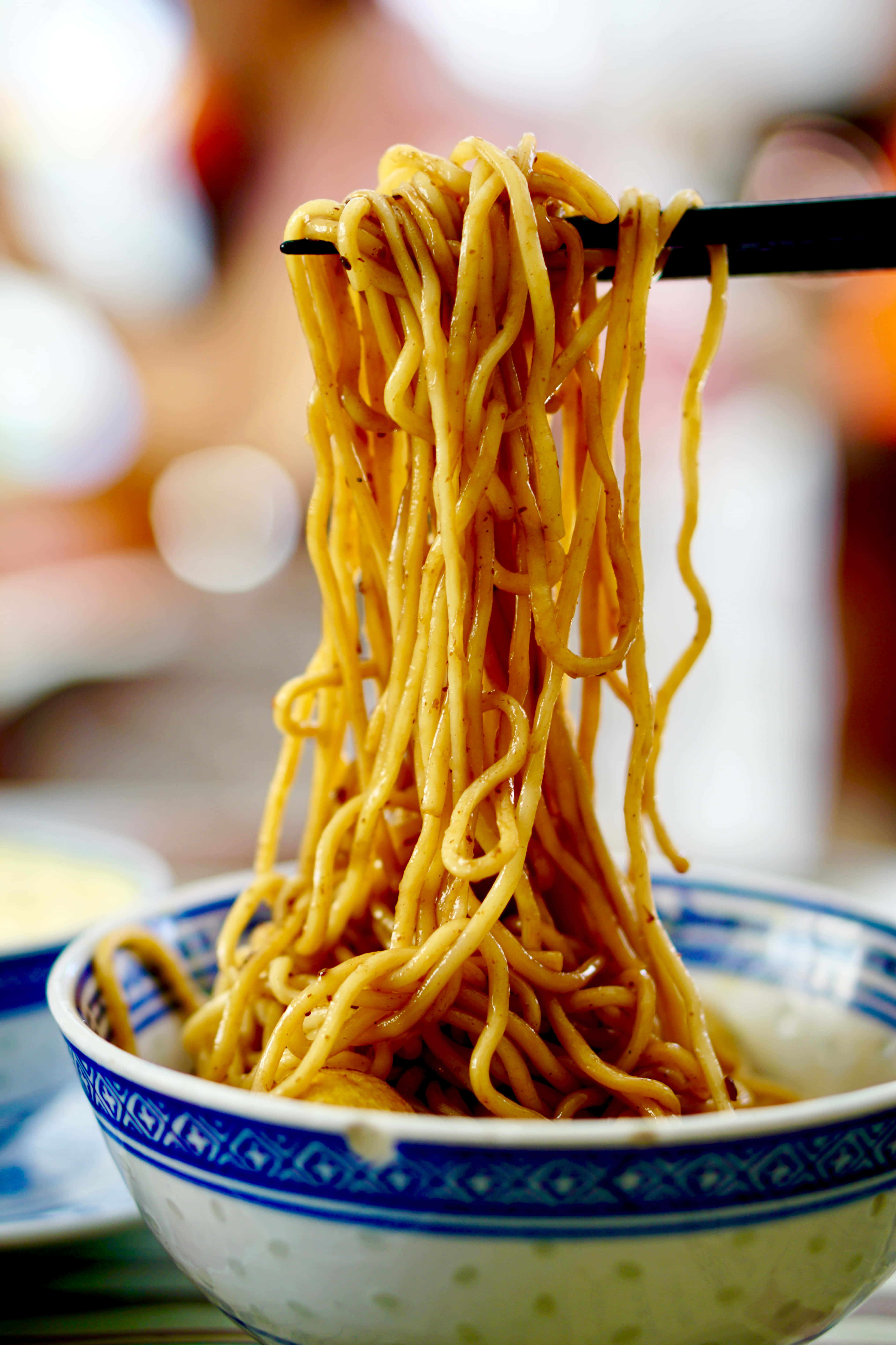 Food Photography 桌布 壁紙 — 黑色筷子從藍白圖案碗中夾起光澤的黃色麵條的特寫。