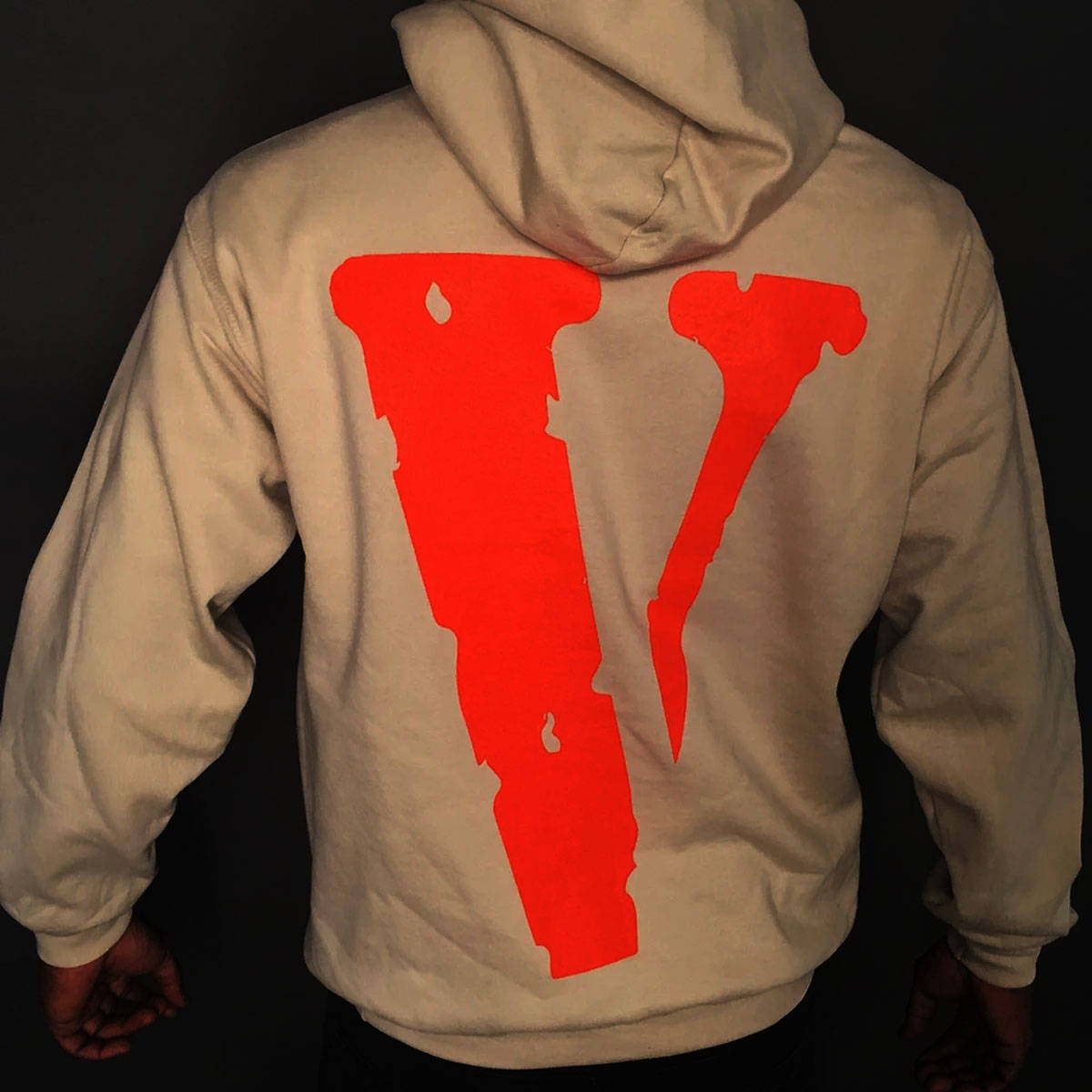 Vlone 桌布 壁紙 — 黑色背景前，一個人穿著米色連帽上衣，背面有一個大膽的紅色風格化V標誌的背面視圖。