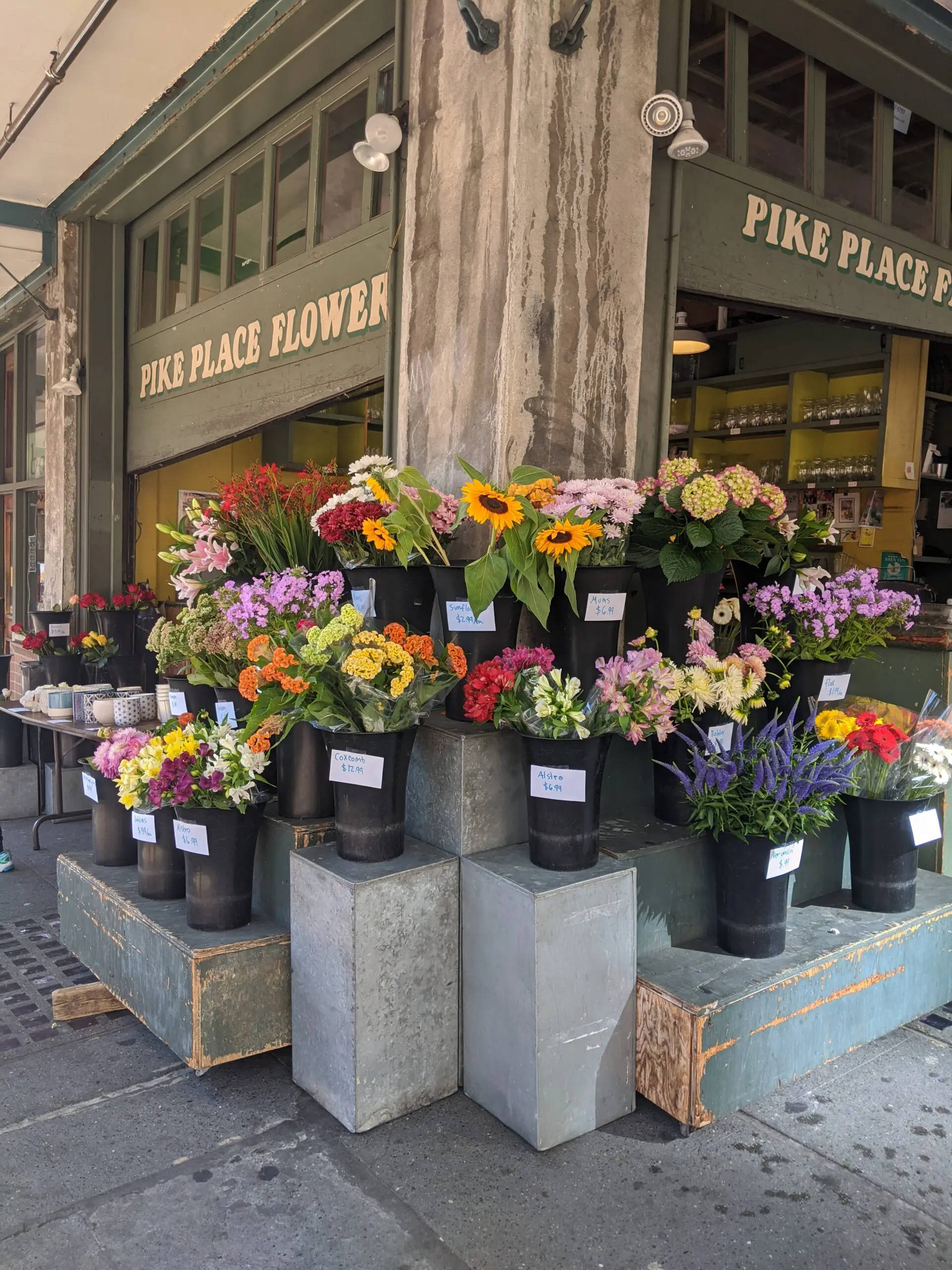 Pike Place Market 桌布 壁紙 — 黑色花盆中充滿活力的花卉佈置，在陽光明媚的城市人行道上，派克市場花店外展示色彩繽紛的花朵。