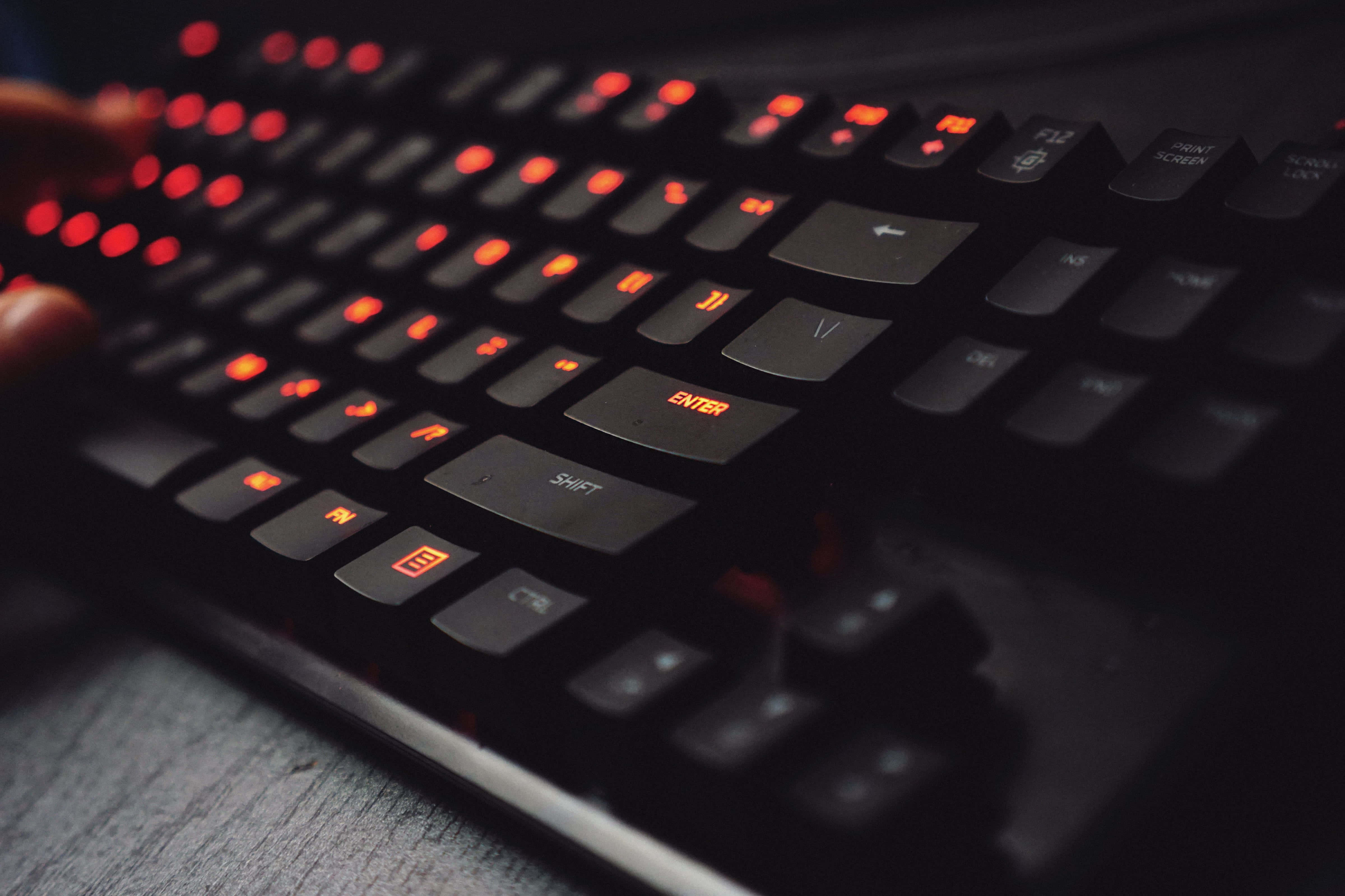 Gaming Keyboards 桌布 壁紙 — 黑色電競鍵盤特寫，帶有發光紅色背光，手指在深色木質表面上打字。