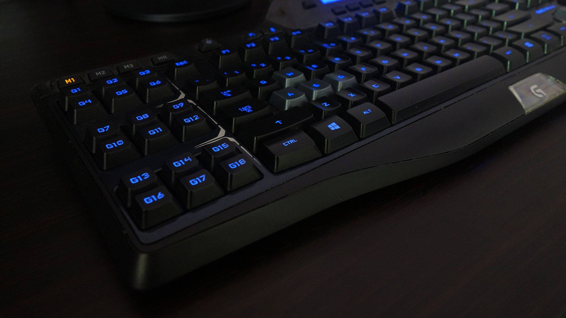 Logitech 桌布 壁紙 — 黑色電競鍵盤特寫，帶有發光藍色背光，放置在昏暗光線下的深色木桌上。