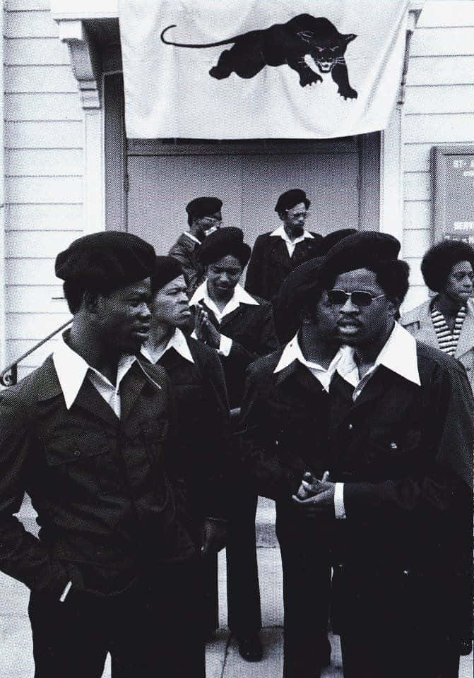 Black Panther Party 桌布 壁紙 — 黑豹黨成員身穿制服，站在一棟建築物外，上方懸掛著黑豹旗幟的黑白照片。