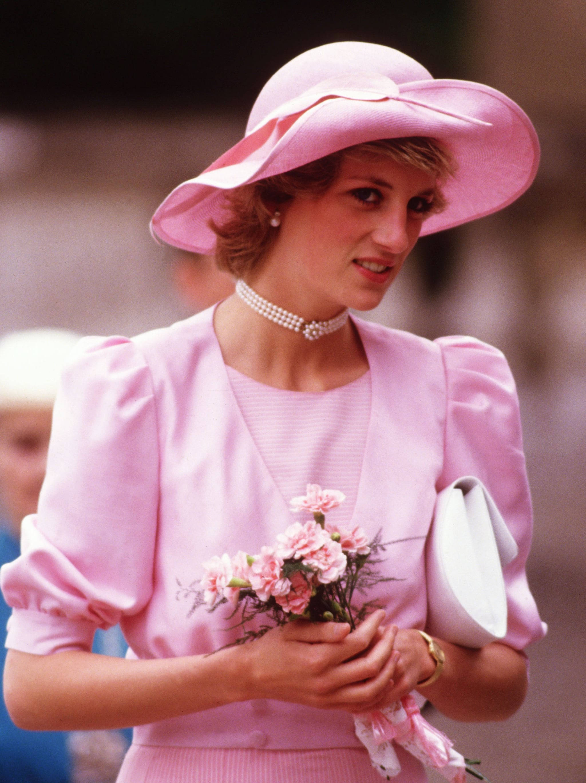 Princess 桌布 壁紙 — 黛安娜王妃身穿柔和粉紅色套裝，搭配相襯帽子和珍珠項鍊，手持粉紅色康乃馨，溫柔微笑。