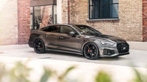 啞光灰色ABT Audi S5 Sportback停在磚砌建築前，前景有模糊的綠色植物。 — 奧迪 GIF 壁紙