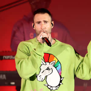 Adam Levine 在舞台上穿著綠色獨角獸毛衣對著麥克風唱歌。 — 1:1 壁紙