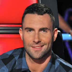 Adam Levine 坐在紅色椅子上微笑，穿著格紋襯衫。 — 1:1 壁紙