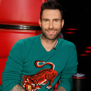 Adam Levine 穿著一件帶有紅色老虎設計的綠色毛衣，在紅色椅子上微笑。 — 1:1 壁紙