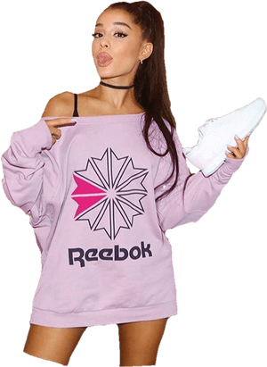 Ariana Grande 身穿淡紫色 Reebok 衛衣，手持白色運動鞋，伸出舌頭，戴黑色頸鏈，馬尾辮。