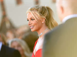 Britney Spears 身穿紅色連衣裙在活動中微笑，背景人群模糊。 — 小甜甜布蘭妮 壁紙