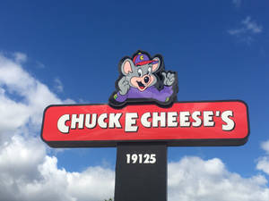 Chuck E Cheese 的吉祥物在藍天白雲下的紅色標誌上揮手。 — 查克芝士 壁紙