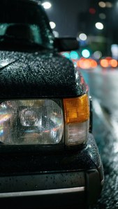 夜晚城市燈光下，黑色汽車引擎蓋上佈滿雨滴。