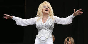 Dolly Parton 在舞台上身穿白色亮片服裝，張開雙臂唱歌。