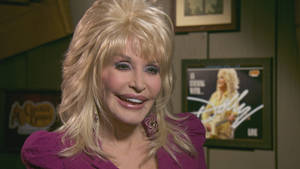 Dolly Parton 身穿紫色上衣微笑，身後有裱框照片。