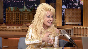 Dolly Parton 身穿金色亮片服裝在脫口秀佈景中微笑。