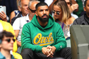 Drake 穿著綠色連帽衫坐在體育館人群中，戴著太陽眼鏡 — MacBook Air 壁紙