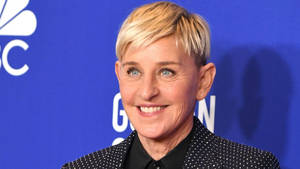 Ellen DeGeneres 在藍色NBC活動背景下穿著黑色圓點西裝外套微笑。