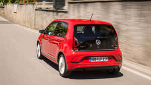 紅色Volkswagen up! TSI在陽光明媚的道路上行駛，旁邊有石牆。 — 福斯汽車 壁紙