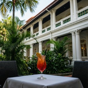 戶外白色桌子上的一杯紅色熱帶飲品，背景有棕櫚樹和殖民建築。