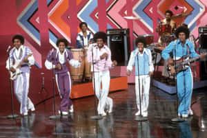 Jackson Five 在帶有幾何圖案和反射地板的色彩繽紛舞台上表演。 — 麥可·傑克森 GIF 壁紙