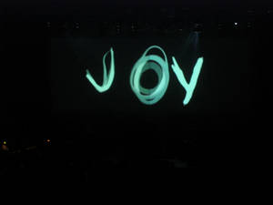 音樂會期間，發光的藍綠色「JOY」字樣投射在黑暗的舞台上。 — 喜悅 壁紙
