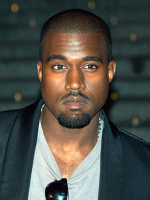 Kanye West 身穿黑色西裝外套和灰色襯衫，太陽眼鏡掛在領口。 — iPad Pro M4 壁紙