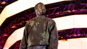 Kanye West 在舞台上穿著深色運動衫和金鍊，在彩色燈光下。 — 肯伊·威斯特 壁紙