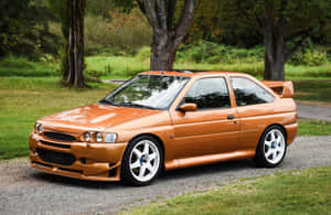 橙色福特 Escort RS Cosworth 停放在樹木和草地附近的碎石上。 — 福特 壁紙