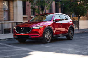 紅色Mazda CX-5 SUV在城市街道上行駛，有磚砌建築和樹木。 — 馬自達 壁紙